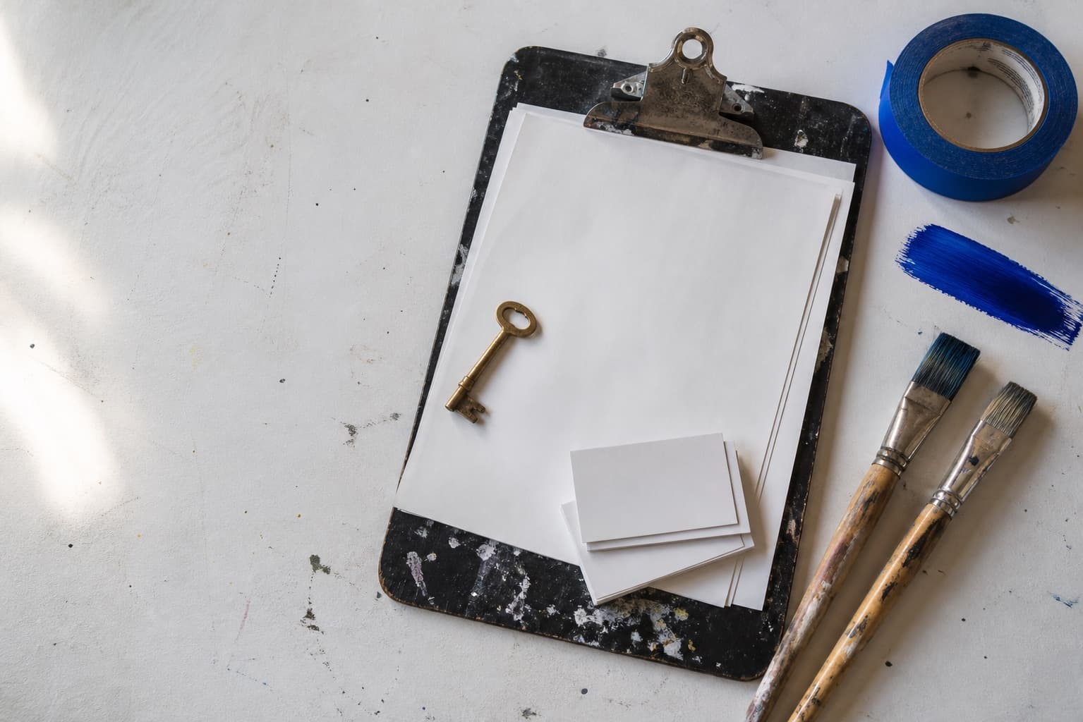 A paint-stained clipboard, studio key, brushes, and blue painter's tape on a worktable